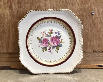 May include: A white square plate with a scalloped edge and a gold and burgundy border. The center of the plate features a floral design with pink roses and green leaves.