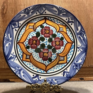 May include: A round ceramic plate featuring a central design of four pink flowers with green leaves surrounded by a yellow geometric border and blue dots. The rim is decorated with blue brushstroke patterns on a white background.