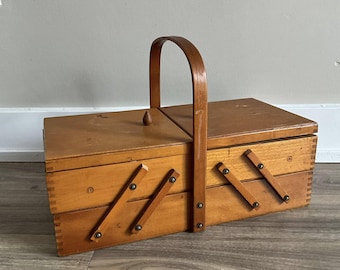 May include: A vintage wooden sewing box with a hinged lid and a curved handle. The box is light brown with visible wood grain and features a series of angled, hinged compartments. The handle is also made of wood.