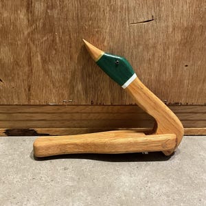 Op de afbeelding: Houten briefopener in de vorm van een eend. De opener heeft een eendenkop met een groen en wit geschilderd ontwerp, een gele snavel en een gebogen houten handvat. De opener is gemaakt van lichtgekleurd hout.