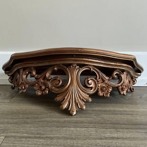 Puede incluir: Estante de pared ornamentado, de color bronce, con una parte superior curvada e intrincados detalles florales y de volutas. El estante está diseñado para montarse en la pared y añade un toque de elegancia vintage. El estante mide aproximadamente 61 cm de ancho.