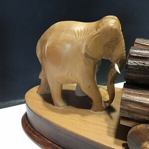 May include: A wooden elephant figurine pulling a cart made of logs. The elephant is light brown and has a white tusk. The cart has two wheels and is made of dark brown wood.
