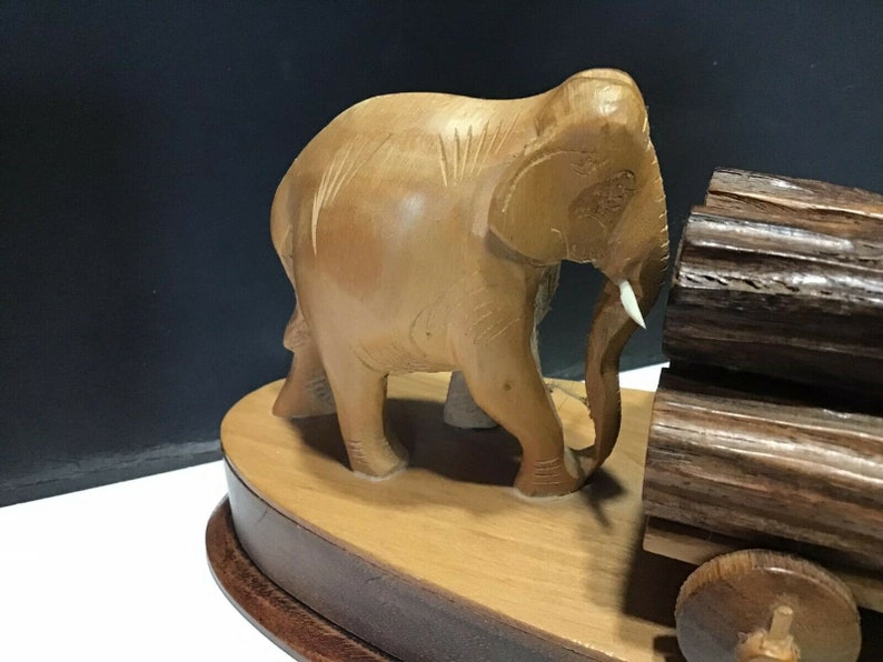 May include: A wooden elephant figurine pulling a cart made of logs. The elephant is light brown and has a white tusk. The cart has two wheels and is made of dark brown wood.