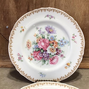 May include: White porcelain dinner plate with a gold rim and a floral design featuring pink, purple, and yellow flowers.