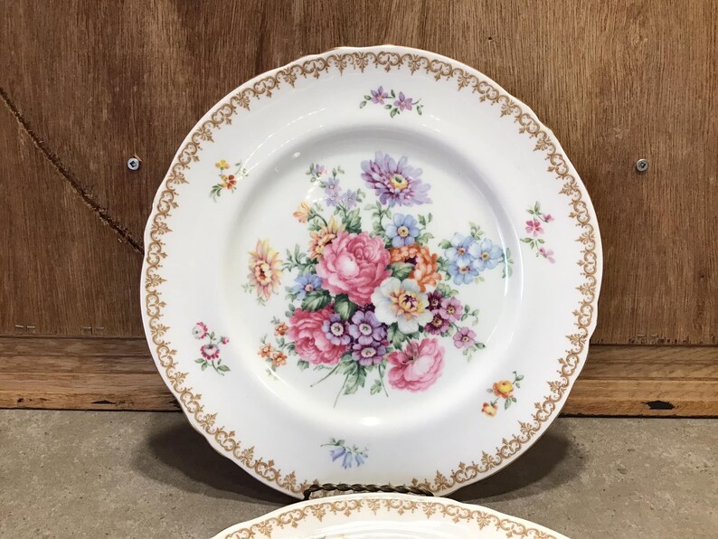 May include: White porcelain dinner plate with a gold rim and a floral design featuring pink, purple, and yellow flowers.