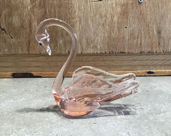 May include: A pink glass swan figurine with a clear glass base. The swan's head is bent down and its wings are spread out.