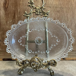 May include: A clear glass oval serving dish with a beaded edge and a decorative pattern of small circles. The dish is displayed on a gold metal stand.