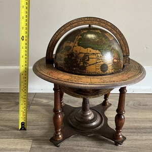 May include: An antique globe on a wooden stand. The globe features a detailed map with green, brown, and tan colors. The stand has a round base with four legs. The globe is approximately 15 inches tall.