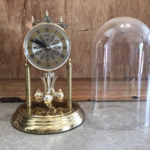 Könnte beinhalten: Eine goldfarbene Pendeluhr mit einer Glaskuppel. Das Zifferblatt der Uhr hat römische Ziffern und ein goldfarbenes Design. Die Uhr steht auf einer Holzoberfläche.