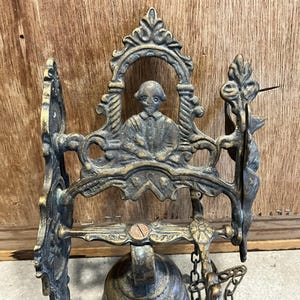 May include: Antique brass bell with an ornate design featuring a figure in a seated pose. The bell is attached to a wall mount and has a chain for ringing.