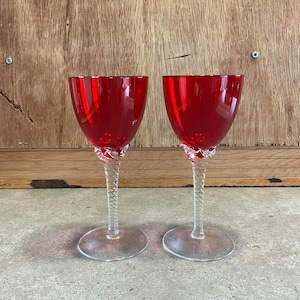 Könnte beinhalten: Zwei rote Weingläser mit gedrehten klaren Stielen und klaren Füßen. Die Gläser stehen auf einer hellbraunen Oberfläche.