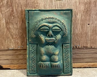 May include: A green ceramic wall plaque with a raised relief sculpture of a stylized human face. The face has closed eyes and a small mouth. The plaque has a textured surface and is decorated with geometric patterns.