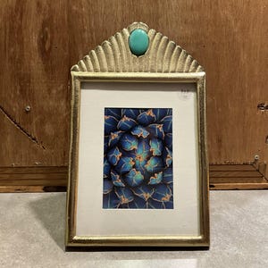 May include: A gold-colored picture frame with a turquoise stone accent at the top. The frame holds a print of a blue and orange floral pattern. The frame is approximately 5x7 inches.