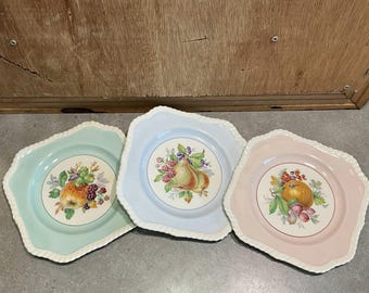 Puede incluir: Tres platos decorativos con bordes festoneados e ilustraciones de frutas. Los platos son de colores pastel: verde menta, azul claro y rosa. Cada plato presenta un arreglo de frutas diferente en el centro, con un borde blanco en forma de cuerda.