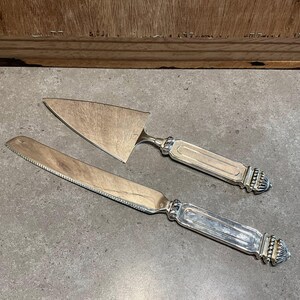 Könnte beinhalten: Silbernes Kuchenservierer- und Messerset. Das Set verfügt über verzierte Griffe mit dekorativen Details. Der Kuchenservierer hat eine dreieckige Klinge, während das Messer eine gezahnte Kante hat. Das Set ist zum Servieren von Kuchen und anderen Desserts bestimmt.