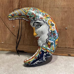 May include: A colorful ceramic crescent moon wall hanging with a painted face and floral designs. The moon is painted in shades of blue, green, yellow, red, and white.