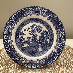 May include: A blue and white ceramic plate with a Willow pattern design. The plate features a detailed landscape scene with a pagoda, trees, and a bridge. The rim has a decorative border. The plate is displayed on a gold-colored metal stand.
