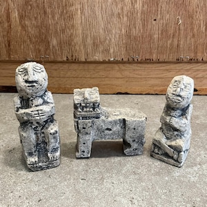 Pode incluir: Três esculturas de pedra desgastada. Duas figuras estão sentadas com as mãos juntas, e uma é um animal estilizado com um corpo retangular e uma face carrancuda. As esculturas são cinzentas com detalhes brancos, sobre um fundo de madeira.
