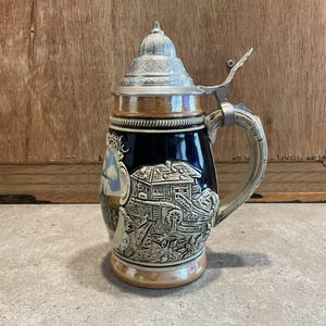May include: A vintage ceramic beer stein with a detailed relief design. The stein has a hinged metal lid, a decorative handle, and a dark blue body with beige and silver trim. The relief depicts a village scene.