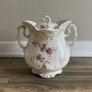 May include: A white porcelain pitcher with a lid, adorned with pink floral patterns and gold accents. The pitcher features two handles and a decorative base. The lid has a small handle. The overall design is ornate and vintage.