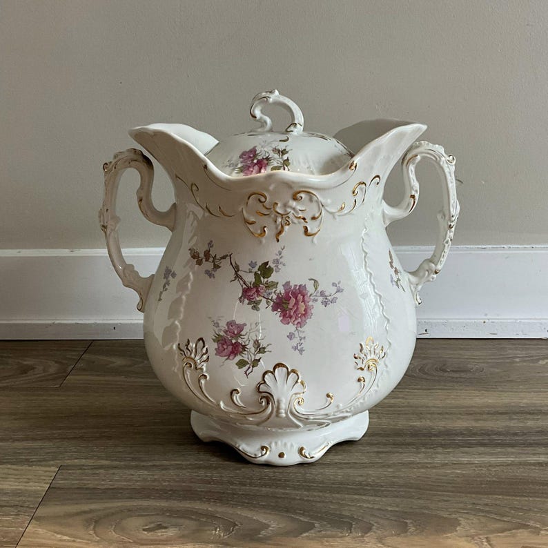 May include: A white porcelain pitcher with a lid, adorned with pink floral patterns and gold accents. The pitcher features two handles and a decorative base. The lid has a small handle. The overall design is ornate and vintage.