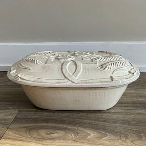 May include: Cream-coloured ceramic casserole dish with a lid. The lid has a raised floral and leaf design with a ribbon detail. The dish has a distressed, aged appearance, and is oval-shaped.