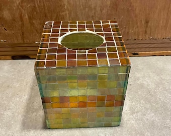 May include: A square tissue box cover made of glass tiles in shades of brown, yellow, and green. The tiles are arranged in a mosaic pattern.