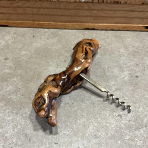 May include: A wooden corkscrew with a natural brown and dark brown wood finish. The corkscrew is made of a single piece of wood with a metal corkscrew attached to the side.