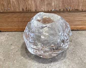 May include: A clear, textured glass votive candle holder. The holder has a rounded shape with a rough, icy appearance. The interior is smooth, designed to hold a small candle. The glass is transparent, allowing light to pass through.