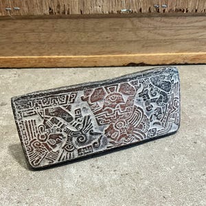 May include: A rectangular, dark brown stone carving with intricate white relief details. The carving depicts stylized figures and geometric patterns, possibly of ancient origin. The stone has a weathered appearance, suggesting age and use.