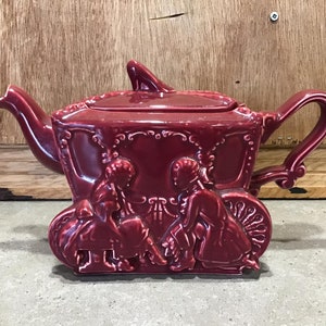 May include: A burgundy ceramic teapot shaped like a horse-drawn carriage with two figures in the front. The teapot has a lid and a handle.