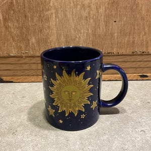 May include: A dark blue ceramic mug with a golden sun design and stars. The mug has a curved handle and is suitable for hot or cold drinks. The celestial theme is prominent. The mug's dimensions are approximately 9 cm in height.
