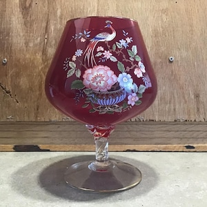 Puede incluir: Una copa de brandy de vidrio rojo rubí con un diseño floral y de pájaros. El diseño presenta un pavo real, flores y follaje en tonos rosa, azul y verde. La base tiene un borde dorado y un tallo retorcido. Altura aproximada: 15 cm.