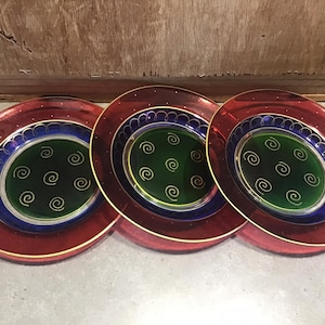 Könnte beinhalten: Drei dekorative Glasteller mit rotem, blauem und goldenem Rand. Die Mitte jedes Tellers zeigt einen grünen Kreis mit einem goldenen Wirbelmuster.