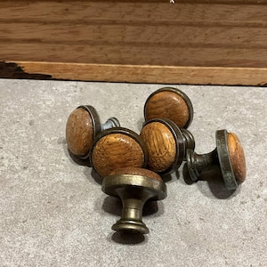 Könnte beinhalten: Eine Sammlung von sechs Vintage-Schrankknöpfen. Die Knöpfe haben eine runde, hellbraune Holzmitte mit einem dunklen Metallrand und -sockel. Die Holzmaserung ist sichtbar. Die Knöpfe sind auf einer grauen Oberfläche angeordnet.