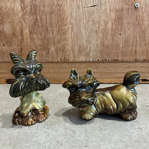 May include: Two ceramic figurines of Scottish Terriers. One is standing with its head tilted and the other is lying down with its head raised. Both are glazed in shades of brown and green.