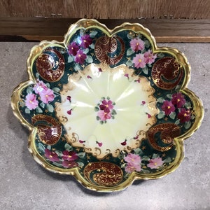 Puede incluir: Un plato decorativo con borde festoneado y diseño floral. El plato presenta un centro amarillo pálido y un borde verde oscuro adornado con flores rosas, patrones de cachemira burdeos y detalles dorados. El borde está adornado con oro.
