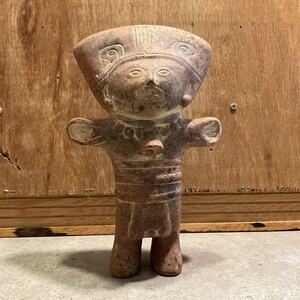 Puede incluir: Figura de terracota de una figura de pie con los brazos extendidos. La figura tiene un tocado ancho y rasgos faciales detallados. El cuerpo está segmentado con un collar y un cinturón detallados. La figura es de color marrón rojizo.