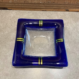 May include: A square glass dish featuring a deep cobalt blue border that transitions into a clear, textured center. The blue rim is accented with four sets of double parallel lime green lines positioned at the center of each side of the square.