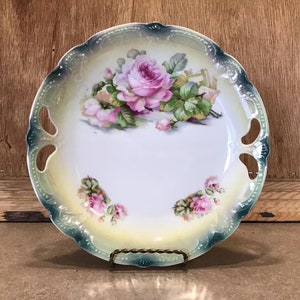 May include: A white porcelain serving plate with a scalloped edge and two cut-out handles. The plate features a floral design with pink roses and green leaves.