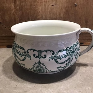 May include: White ceramic chamber pot with a green and white floral design. The pot has a wide, round base and a handle on the side.