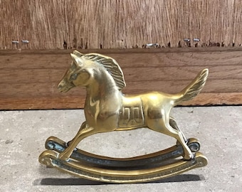 Puede incluir: Una figurita de caballo de balancín de latón de color dorado. El caballo está en posición de galope con la cabeza girada hacia un lado. La base de balancín está hecha de dos piezas de latón curvadas.