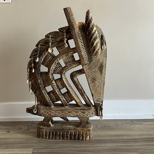 Puede incluir: Escultura de madera tallada de una cabeza de caballo. La escultura marrón presenta intrincados patrones y una estructura similar a una melena. Pequeñas conchas están unidas a la melena. La cabeza de caballo está montada sobre una base rectangular.