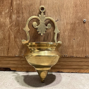 May include: A vintage brass wall sconce with a decorative floral design and a rounded bowl shape. The sconce has a loop at the top for hanging and a pointed bottom. The brass has a polished finish.