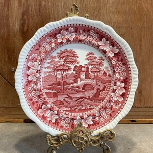 May include: A red and white ceramic plate with a floral design and a scene of a bridge and trees. The plate is on a gold metal stand.