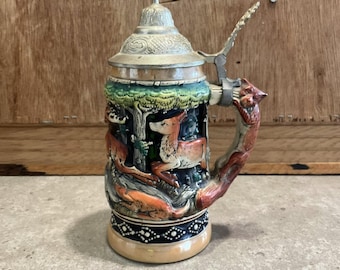 Könnte beinhalten: Ein Vintage-Bierkrug mit einer detaillierten Waldszene mit Hirschen und Füchsen. Der Keramikkörper hat einen dekorativen Sockel und einen Metalldeckel mit einem Klappgriff. Der Griff ist wie ein Fuchs geformt.