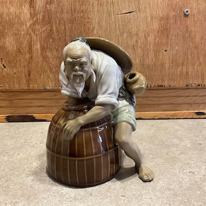 May include: A ceramic figurine of a man with a white beard and a straw hat, holding a brown barrel. The figurine is wearing a white shirt and green trousers.