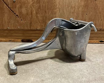 May include: A vintage, silver-colored citrus juicer. The juicer has a lever arm, a conical reamer, and a juice collection cup. The base has three small feet. The juicer is made of metal and appears to be well-used.