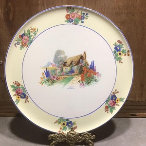May include: A white ceramic plate with a yellow rim and blue and floral accents. The centre of the plate features a painted image of a cottage with a thatched roof and a garden.