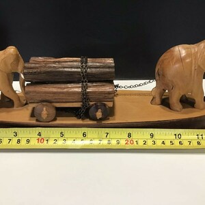 May include: Wooden figurine of two elephants pulling a cart with logs. The cart has wheels and a chain connecting the elephants to the cart. The figurine is on a wooden base.